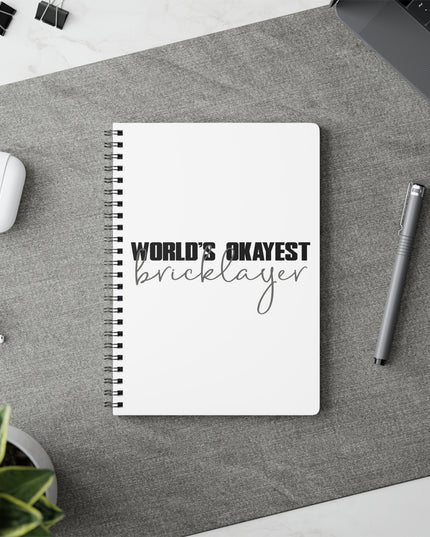 Notebook with 'World's Okayest Bricklayer' text on a desk with a pen and earbuds.