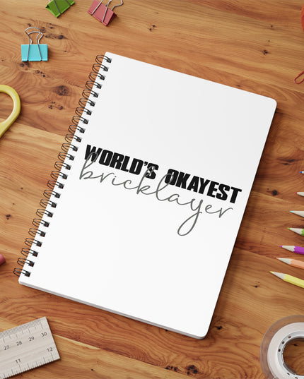 Notebook with 'World's Okayest Bricklayer' text on a wooden surface with stationery items.