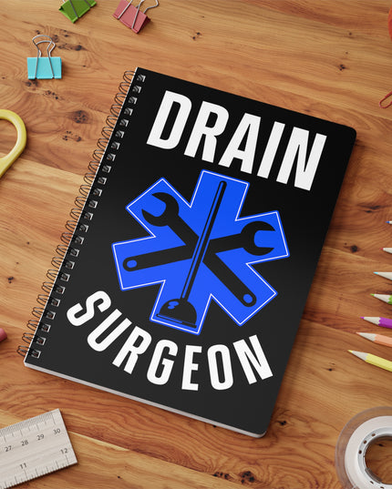 Black spiral A5 plumber notebook with "Drain Surgeon" text and a blue medical cross graphic that include plumbing tools, on a wooden table