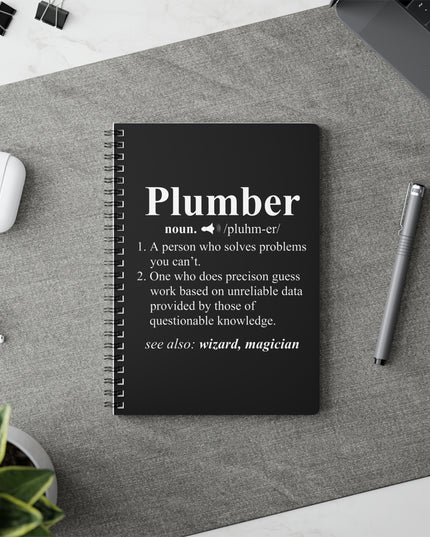 Black notebook with 'Plumber' definition on a gray surface with a pen and airpods.