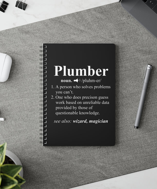 Black notebook with 'Plumber' definition on a gray surface with a pen and airpods.